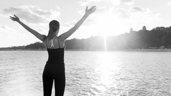 Frau im Sportdress stehend von hinten mit hochgestreckten Armen genießt am Wasser das Sonnenlicht.